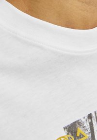 Hvid bomulds T-shirt med rund hals, der har et trykt design i grå og gul på forsiden, der viser geometriske og naturlige elementer.