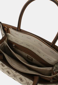 Grand sac fourre-tout avec un extérieur synthétique marron, une doublure intérieure beige imprimée du mot « GUESS », plusieurs compartiments et une fermeture éclair.
