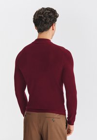 Bordowy sweter dzianinowy z długimi rękawami i okrągłym dekoltem, o gładkiej fakturze, pokazany od tyłu. Widoczne brązowe spodnie.