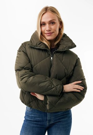Blonde Frau trägt eine olivgrüne gesteppte Steppjacke mit hohem Kragen und blaue Jeans, verschränkte Arme und leicht lächelnd.