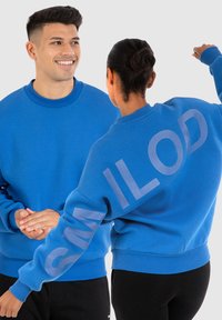 Felpe blu realizzate in morbido tessuto, con scollo rotondo e vestibilità oversize. Una di esse presenta la parola "SMILOD" stampata sulla manica in azzurro chiaro.