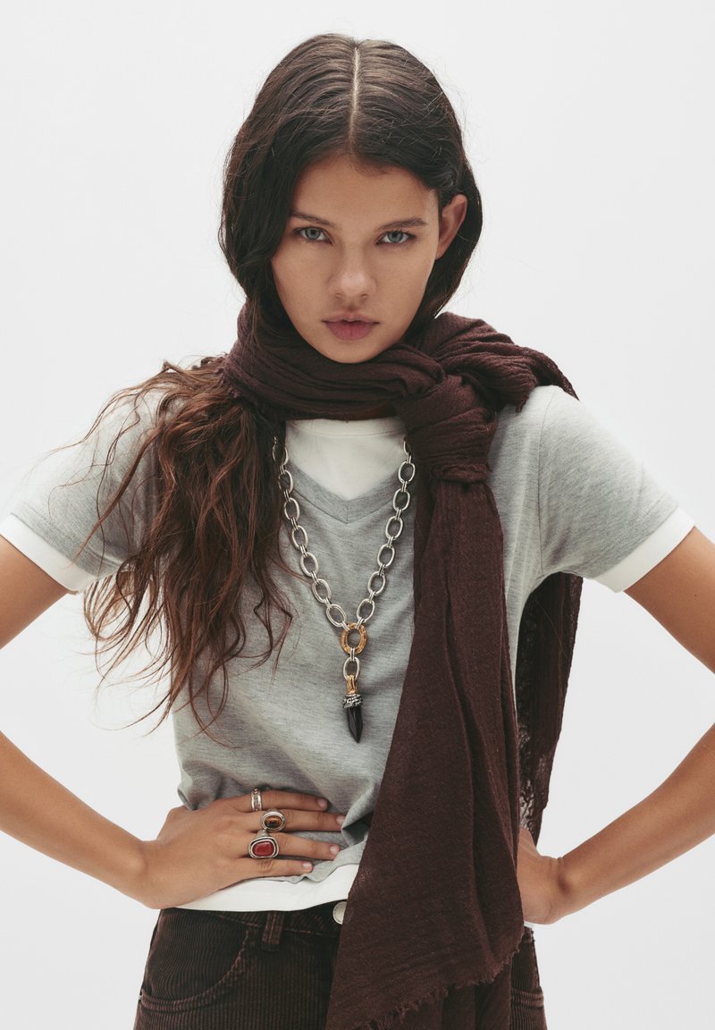 T-shirt grigio a scollo a V sovrapposto a una maglietta bianca, abbinato a una sciarpa marrone scuro. Gli accessori comprendono una collana in catena spessa e diversi anelli.