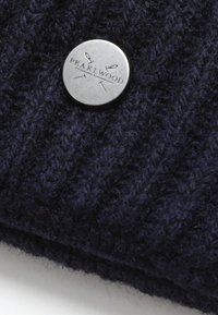 Tissu tricoté bleu marine avec une texture côtelée. Présente un bouton en métal rond embossé avec "PEARLWOOD" et des motifs de ciseaux.