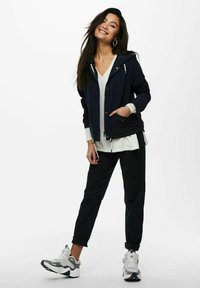 Navy jacket with hood and zipper, layered over a white top. Black pants and gray sneakers with white accents complete the outfit.