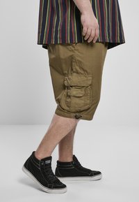 Khaki šortky v olivovo zelenej farbe, s dĺžkou po kolená a bočnými vreckami, skombinované s čiernymi vysokými teniskami a pruhovanou košeľou v niekoľkých farbách.