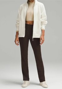 Personne portant une veste blanche zippée, un crop top beige, un pantalon marron foncé et des baskets blanches, debout devant un fond uni.