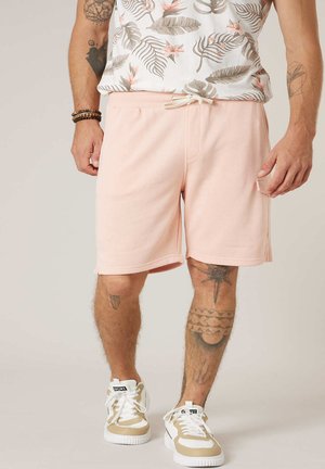 Homme portant un short à cordon rose, une chemise sans manches à motifs floraux, des baskets blanches et beiges, et des bracelets, montrant des tatouages sur la jambe et le bras.