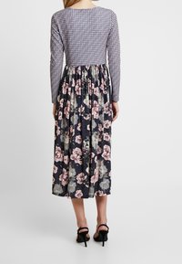 Vrouw in een jurk met lange mouwen, een gebloemde bodice en een rok met bloemenprint, gecombineerd met zwarte hoge hakken, staande met haar rug naar de camera.