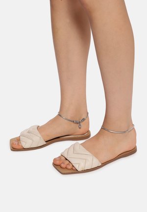 Lysebeige flade sandaler med et vævet stropdesign, firkantet tå og tynd sål. Anklet tilføjer detaljer til præsentationen.