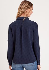 Mørkeblå langærmet skjorte med et subtile tekstur mønster, krave, knap bind og en afslappet pasform. Har en yoke og plissé detalje.