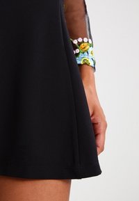 Svart klänning med plisserad nederkant, som har ett texturerat tyg. Arm prydd med ett färgglatt broderat armband med blommönster.