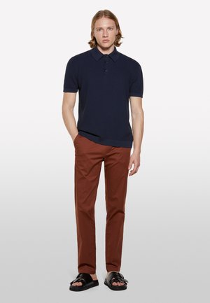 Polo à manches courtes bleu marine, en tissu texturé, avec patte de boutonnage à trois boutons. Associé à un pantalon rouille et des sandales noires avec une boucle.