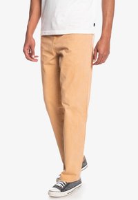 Pantaloni di cotone beige con un design a gamba dritta, caratterizzati da una texture liscia e dettagli cuciti, abbinati a sneakers grigie.