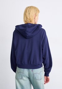 Persona con cabello corto rubio, vistiendo una sudadera azul marino y jeans azul claro, de espaldas frente a un fondo blanco liso.