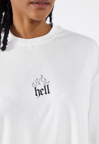 Hvid bomulds T-shirt med et lille sort flamme-design over ordet "hell" i stiliseret skrifttype, rund halsudskæring og afslappet pasform.