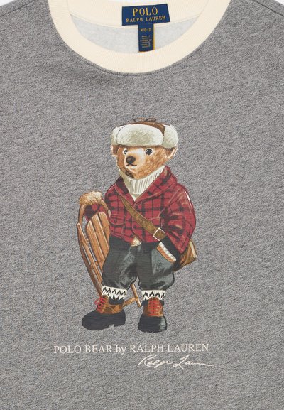 Pelēks džemperis ar lāča ilustrāciju, kam mugurā sarkans čekots krekls, balta šalle un ziemas cepure, kurš tur slēpošanas kamanas. Zem attēla ir teksts "POLO BEAR by RALPH LAUREN."
