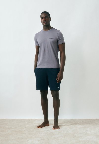 Νέος άνδρας που στέκεται ξυπόλυτος πάνω σε χαλί, φορώντας γκρι εφαρμοστό μπλουζάκι και σκούρα μπλε σορτς, μπροστά σε απλό λευκό τοίχο.