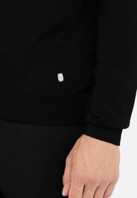 Pulover negru tricotat, cu margine și manșete cu carouri, având un tag mic cu logo argintiu pe partea inferioară. Textură moale, design simplu.