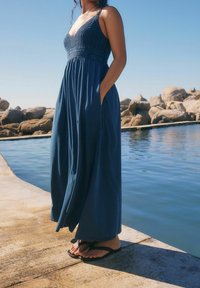 Robe longue bleue foncée sans manches avec un corsage texturé, une jupe fluide et des poches. Portée avec des tongs noires, debout près de l'eau.
