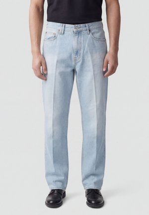 Uomo che indossa jeans celesti a gamba dritta, scarpe nere e camicia nera, in piedi davanti a uno sfondo chiaro e semplice.