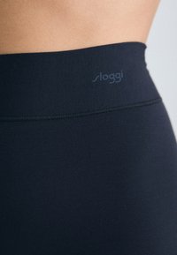 Crne tajice s glatkom teksturom, elastičnim pojasom koji sadrži suptilan "sloggi" logotip, te uskim dizajnom koji naglašava udobnost i fleksibilnost.