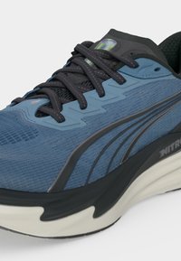 Basket de sport bleu et noir avec une tige en tissu texturé, des lacets noirs, une semelle épaisse blanche et noire, et des détails de marque sur le côté et la languette.