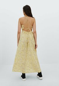 Robe longue jaune avec un motif floral texturé, bretelles halter, dos ouvert, taille froncée, associée à des baskets noires.