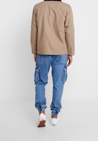 Henkilö, joka on pukeutunut beigeen takkiin, sinisiin denim-sovittimiin elastisilla hihansuilla ja valkoisiin tossuihin, seisoo yksinkertaista valkoista taustaa vasten.