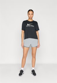 Μαύρο cropped t-shirt Nike Pro με λευκό λογότυπο, συνδυασμένο με ανοιχτό γκρι σορτς. Το μοντέλο φοράει μαύρα αθλητικά παπούτσια με λευκές λεπτομέρειες. Απλό φόντο.