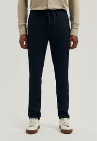 Man in een beige shirt met lange mouwen, marineblauwe joggingbroek met trekkoord en witte sneakers, staand voor een effen achtergrond.
