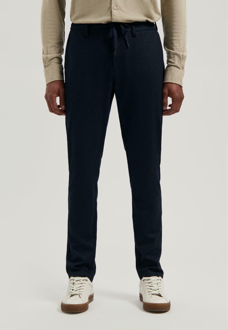 Man in een beige shirt met lange mouwen, marineblauwe joggingbroek met trekkoord en witte sneakers, staand voor een effen achtergrond.