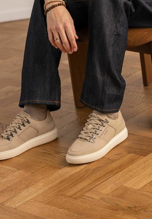 Persoon zittend op een houten stoel, gekleed in donkere jeans, beige sneakers met metalen oogjes, beige sokken en armbanden aan de linker pols.