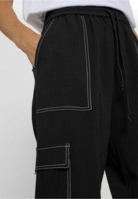 Pantalon cargo noir en tissu texturé, avec surpiqûres blanches, taille à cordon de serrage et poches latérales pour le rangement.