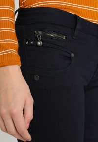Jeans azul marino con un bolsillo con cremallera y herrajes metálicos, tela texturizada y costuras visibles. La mano descansa cerca del bolsillo.