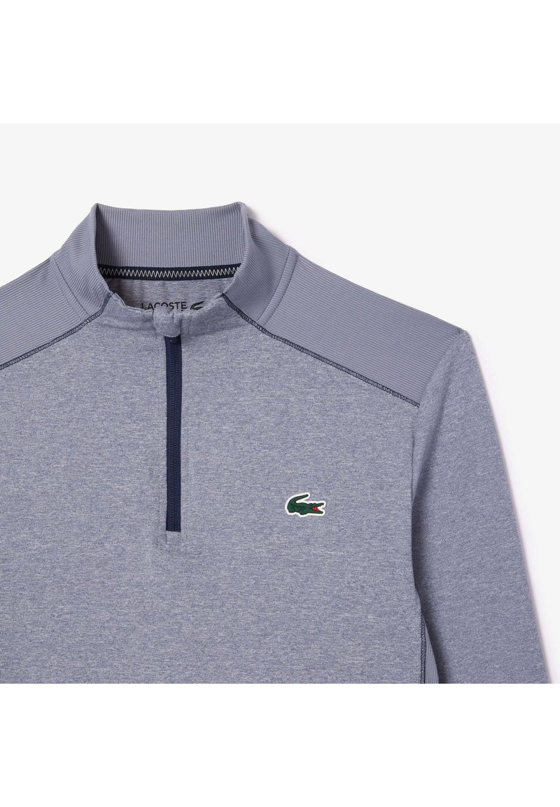 Hellgrauer Pullover mit langen Ärmeln und Reißverschluss am Hals. Gerippter Kragen und Schulterakzente. Mit einem grünen Krokodil-Logo auf der Brust.