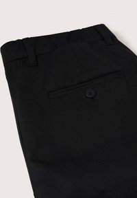 Gros plan sur un pantalon noir sur mesure montrant la taille, les passants de ceinture et une poche arrière avec bouton sur un fond clair.