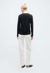 Άτομο με κοντά ξανθά μαλλιά που κοιτάζει προς τα πίσω, φοράει μαύρο μακρυμάνικο πουκάμισο, άσπρο παντελόνι και μαύρα παπούτσια με τακούνι, στέκεται σε απλό φόντο.