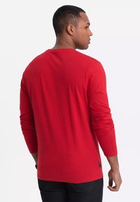 Punainen pitkähihainen t-paita, joka on valmistettu puuvillasta, pyöreällä kaula-aukolla ja suoran helman kanssa. Siinä on pieni brändiläppä alaosassa.