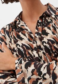 Camicia abbottonata color beige con un motivo astratto in marrone e nero, caratterizzata da pieghe verticali, colletto e maniche lunghe.