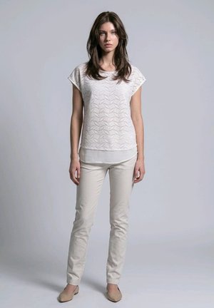 Mujer joven con cabello oscuro que lleva una camiseta blanca texturizada de manga corta, pantalones beige claro y zapatos planos beige, de pie contra un fondo liso.