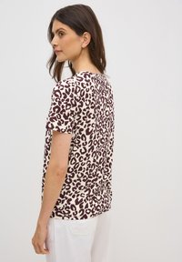 Camiseta de manga corta en color crema con un estampado de animales en burdeos, con cuello redondo y un ajuste relajado. Pantalones blancos al fondo.