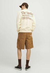 Sweatshirt crem cu text maro pe spate, pantaloni scurți maro tip cargo, șosete maro și adidași negri cu talpă albă. O ținută casual, cu un design minimalist.