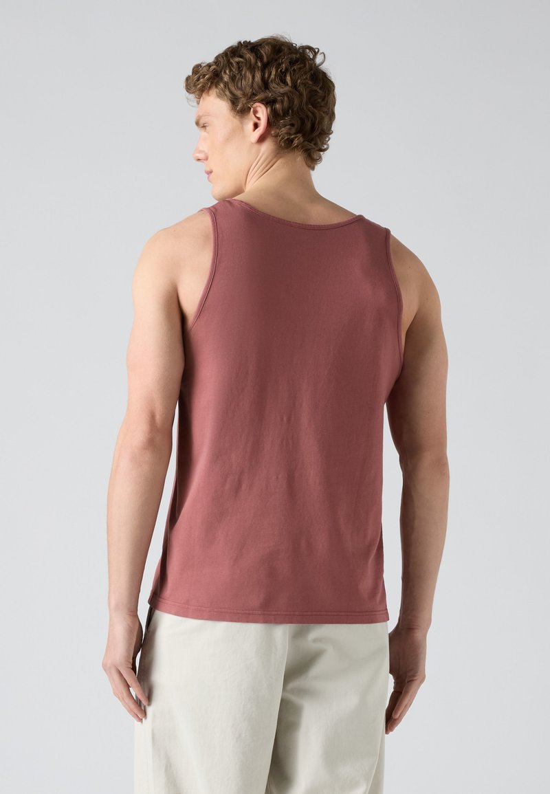 Mann mit lockigem Haar, der ein ärmelloses, staubrosafarbenes Tanktop und hellbeige Hosen trägt, von hinten vor einem einfarbigen grauen Hintergrund gezeigt.