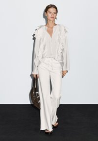 White ruffled blouse with buttons, paired with tailored white high-waisted pants. Accessory: dark brown handbag. Smooth textures, modern design.