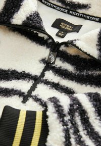 Fleece halvlynjakke med sort-hvide zigzag-striber. Tekstureret stof med ribbede manchetter, fremhævet af gule striber. Brandmærke synligt.