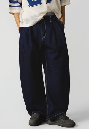 Person iført løse mørkeblå vidde-ud bukser med hvide kontrastsømme, sorte sneakers og en hvid mesh-top med blå taltryk.