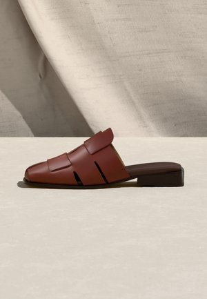 Ruskeat nahkaiset slip-on-mule-kengät, joissa on leikattuja hihnoja ja matala paksukorko, valkoisella teksturoidulla pinnalla ja kangastaustalla.