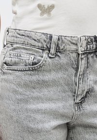 Γκρι ψηλόμεσα τζιν με όξινη πλύση, κλείσιμο με κουμπιά και μικρή μπροστινή τσέπη, συνδυασμένα με λευκό τοπ που διαθέτει διακοσμητικό πεταλούδα από κρύσταλλα.