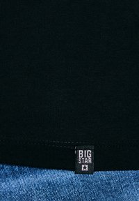 Mustast kangast, mille serva lähedale on tikitud "BIG STAR" brändisilt, allpool sinise teksariide kangaga.