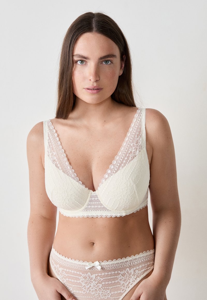Giovane donna con lunghi capelli castani che indossa un reggiseno bianco in pizzo e mutandine abbinate in pizzo su uno sfondo chiaro uniforme.